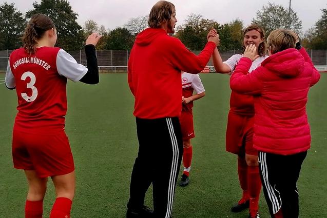 Eine Gruppe von Personen in roten Fu&szlig;balltrikots feiert auf einem Kunstrasenplatz, umgeben von B&auml;umen und einem Schiedsrichter.