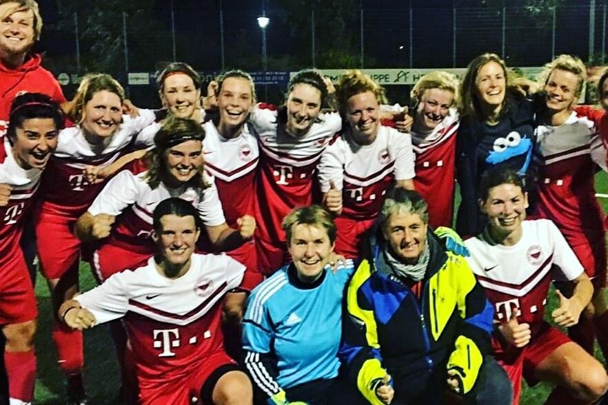 Gruppenselfie von Frauen in roten Fu&szlig;balltrikots, umgeben von Trainer und Betreuern auf einem Kunstrasenplatz.