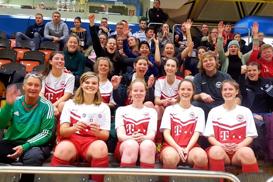 Gruppenshot von feiernden Fu&szlig;ballspielerinnen und Zuschauern auf einer Trib&uuml;ne, alle winken und zeigen Freude.