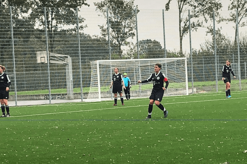 Spielerinnen in schwarzen Trikots auf einem Kunstrasenfeld, im Hintergrund ein Fu&szlig;balltor und eine Torh&uuml;terin in Blau.