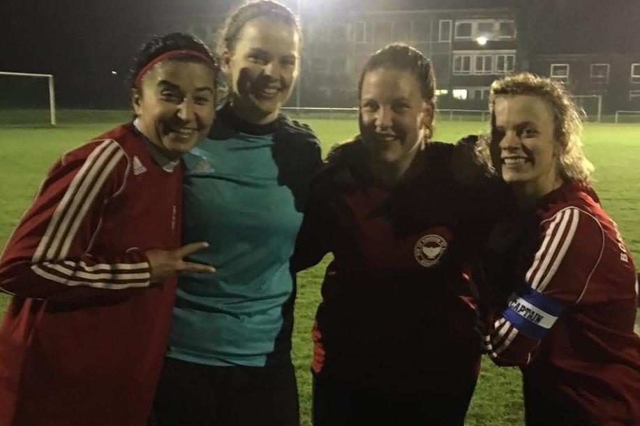 Vier Frauen in Sportkleidung stehen l&auml;chelnd auf einem Fu&szlig;ballfeld bei Nacht, begrenzt von nur schwachem Licht.