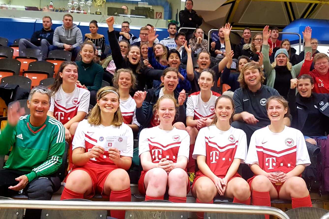 Gruppenshot von feiernden Fu&szlig;ballspielerinnen und Zuschauern auf einer Trib&uuml;ne, alle winken und zeigen Freude.