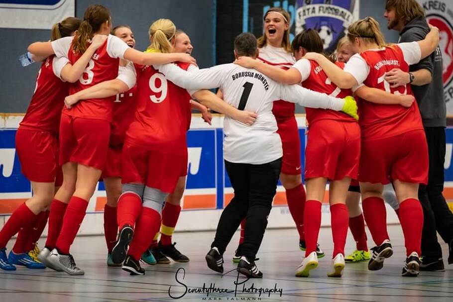 Mannschaft von Frauen im roten Trikot umarmt sich freudig in einer Sporthalle vor einer Wand mit Vereinswappen.