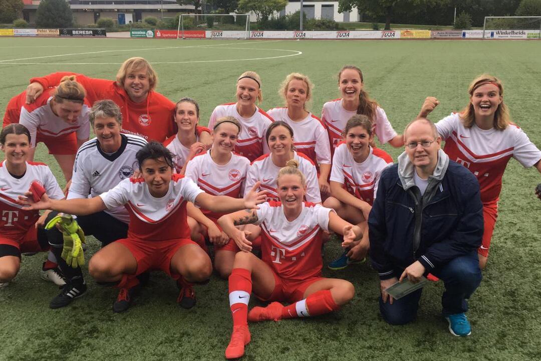 Gruppenshooting von Fu&szlig;ballspielerinnen in roten Trikots auf dem Sportplatz, umgeben von Trainer und Betreuern.