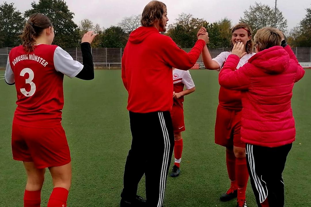 Eine Gruppe von Personen in roten Fu&szlig;balltrikots feiert auf einem Kunstrasenplatz, umgeben von B&auml;umen und einem Schiedsrichter.