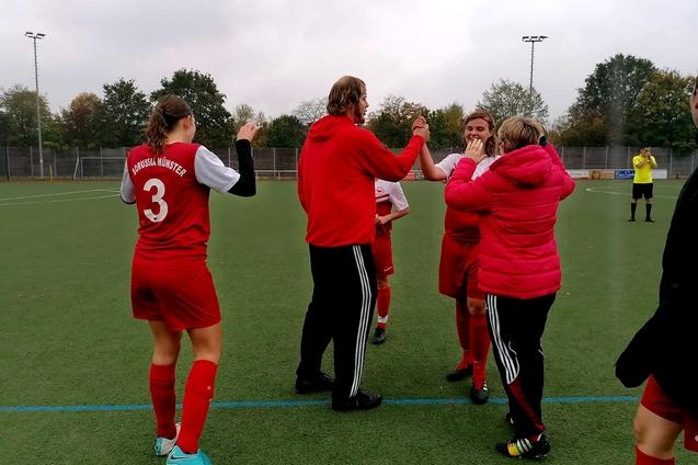 Spielerinnen und Trainer feiern einen Torerfolg auf dem Fu&szlig;ballfeld, umgeben von gr&uuml;ner Rasenfl&auml;che und B&auml;umen im Hintergrund.