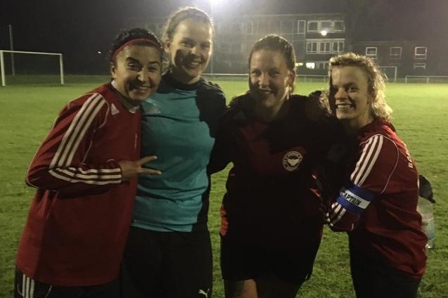 Vier Frauen in Sportkleidung stehen l&auml;chelnd auf einem Fu&szlig;ballfeld bei Nacht, begrenzt von nur schwachem Licht.