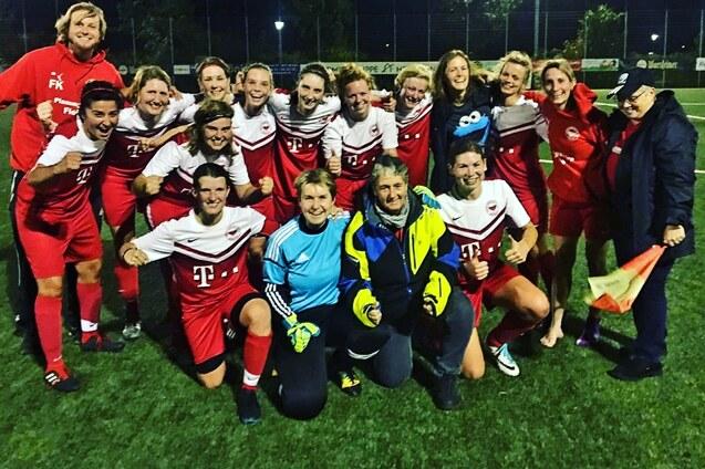 Gruppenselfie von Frauen in roten Fu&szlig;balltrikots, umgeben von Trainer und Betreuern auf einem Kunstrasenplatz.