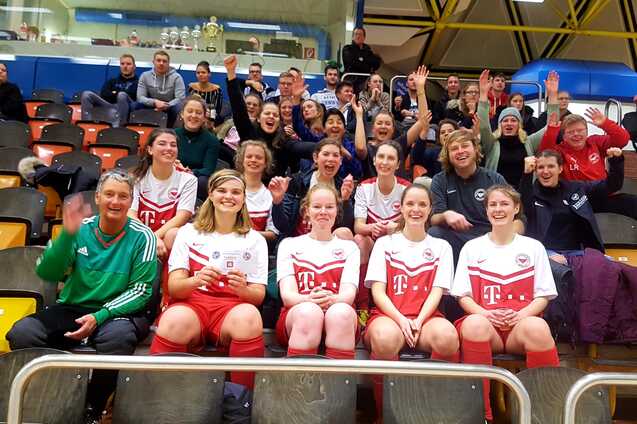 Gruppenshot von feiernden Fu&szlig;ballspielerinnen und Zuschauern auf einer Trib&uuml;ne, alle winken und zeigen Freude.