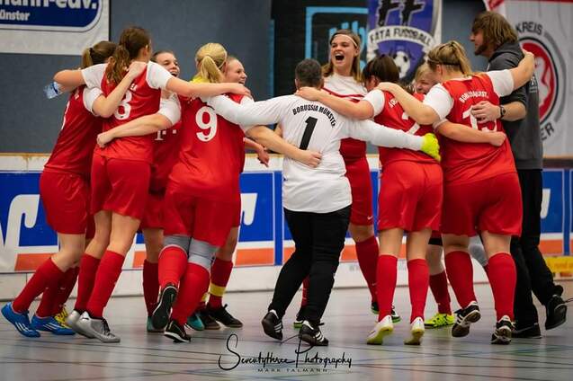 Mannschaft von Frauen im roten Trikot umarmt sich freudig in einer Sporthalle vor einer Wand mit Vereinswappen.