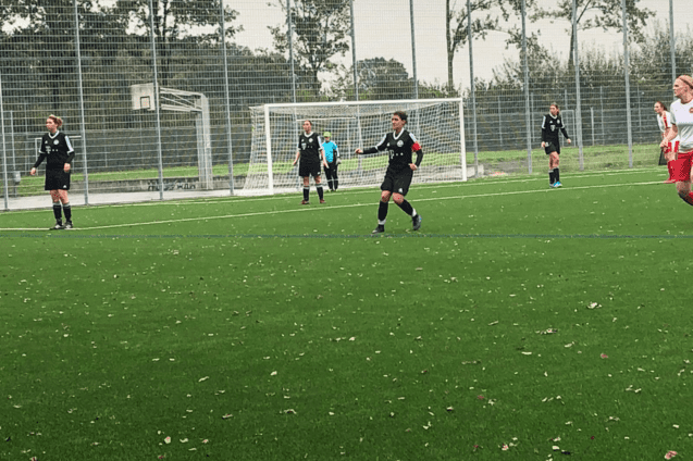Spielerinnen in schwarzen Trikots auf einem Kunstrasenfeld, im Hintergrund ein Fu&szlig;balltor und eine Torh&uuml;terin in Blau.