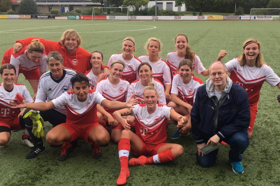 Gruppenshooting von Fu&szlig;ballspielerinnen in roten Trikots auf dem Sportplatz, umgeben von Trainer und Betreuern.