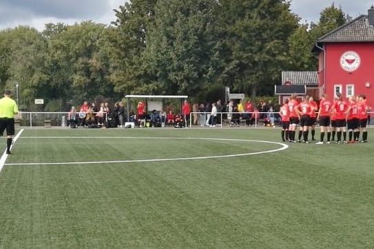 Sportplatz mit Kunstrasen, einer Fu&szlig;ballmannschaft in roten Trikots, einem Schiedsrichter und Zuschauer im Hintergrund.