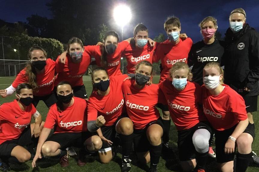 Gruppenshot von 13 Frauen im Fu&szlig;balltrikot mit Masken, stehend und kniend auf einem Fu&szlig;ballfeld bei Abendd&auml;mmerung.
