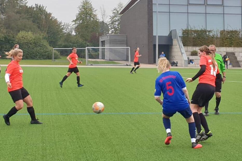 Fu&szlig;ballspiel auf Kunstrasen, mit Spielerinnen in roten Trikots, einer mit der Nummer 5 und einer in Blau, im Ballbesitz.