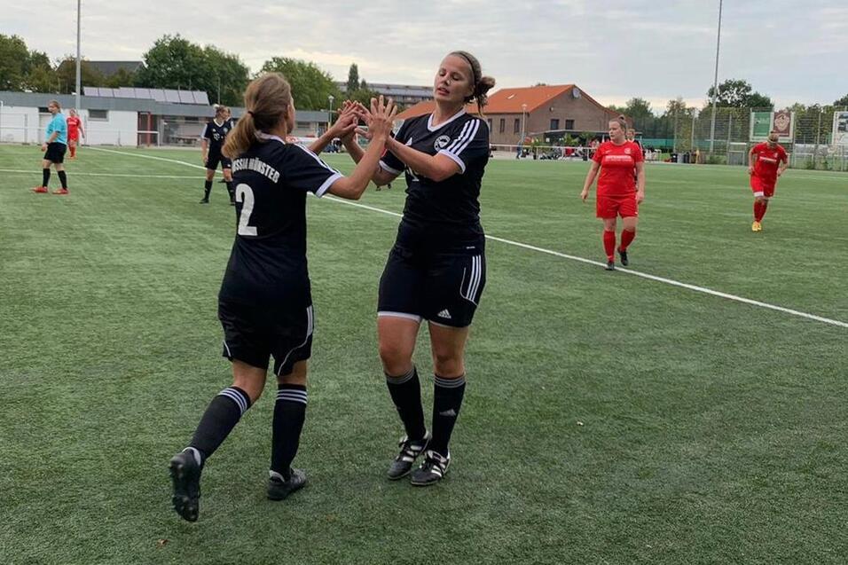 Zwei weibliche Fu&szlig;ballspielerinnen in schwarzen Trikots feiern auf einem Kunstrasenplatz, w&auml;hrend andere Spieler im Hintergrund agieren.