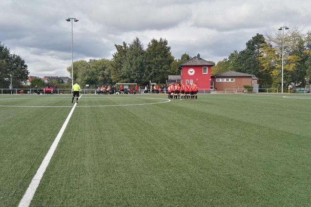 Sportplatz mit Kunstrasen, einer Fu&szlig;ballmannschaft in roten Trikots, einem Schiedsrichter und Zuschauer im Hintergrund.