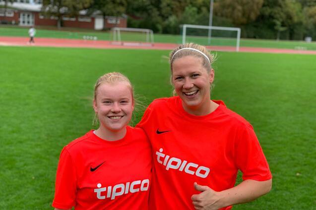 Zwei Fu&szlig;ballspielerinnen in roten Trikots mit "tipico"-Aufdruck l&auml;cheln auf einem Sportplatz.