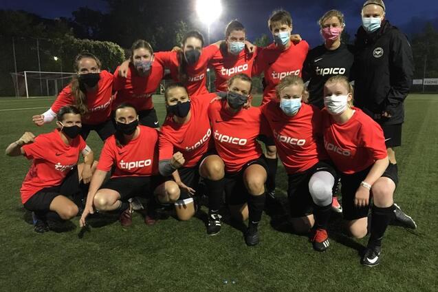 Gruppenshot von 13 Frauen im Fu&szlig;balltrikot mit Masken, stehend und kniend auf einem Fu&szlig;ballfeld bei Abendd&auml;mmerung.
