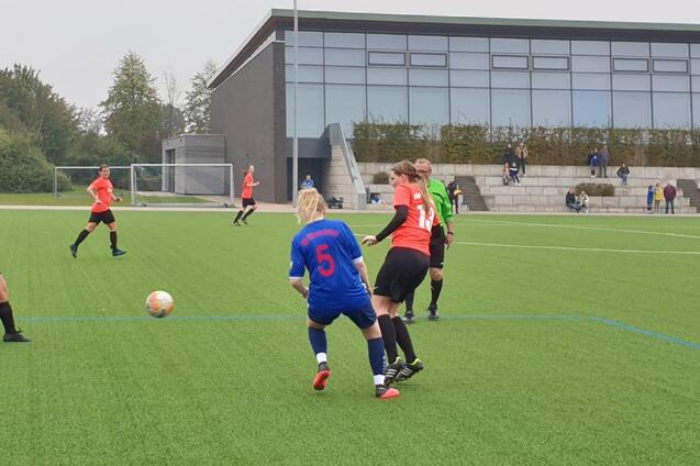 Fu&szlig;ballspiel auf Kunstrasen, mit Spielerinnen in roten Trikots, einer mit der Nummer 5 und einer in Blau, im Ballbesitz.