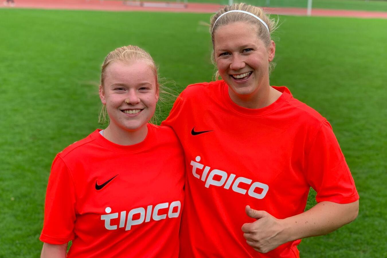 Zwei Fu&szlig;ballspielerinnen in roten Trikots mit "tipico"-Aufdruck l&auml;cheln auf einem Sportplatz.