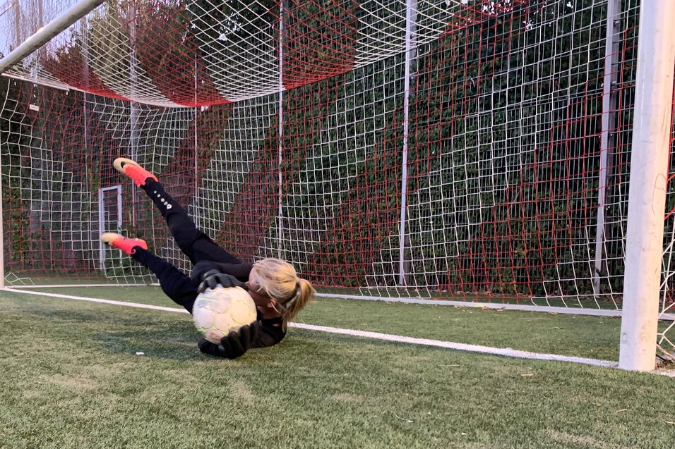 M&auml;dchen in der Torwartposition f&auml;ngt einen Fu&szlig;ball vor einem Tor auf einem Kunstrasenplatz.