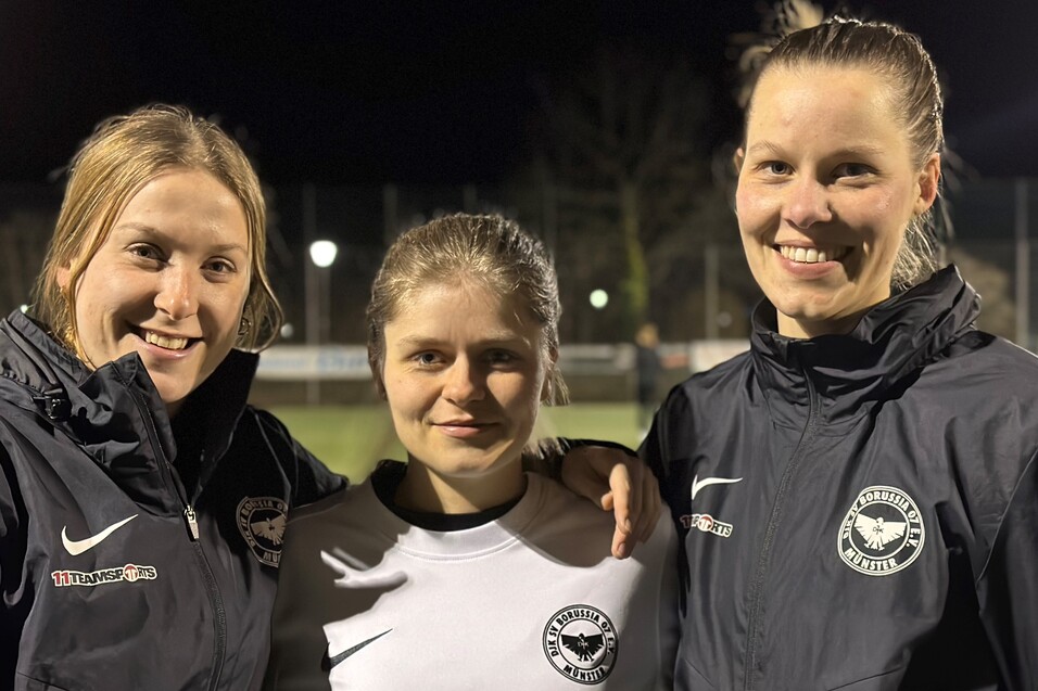 Drei Frauen in Sportjacken stehen l&auml;chelnd auf einem Fu&szlig;ballplatz bei Nacht, eine tr&auml;gt ein wei&szlig;es Trikot.