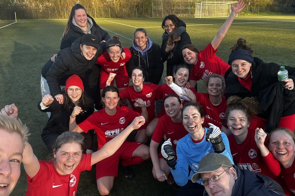Gruppenselfie von Frauen in roten Fu&szlig;balltrikots auf einem Feld, alle l&auml;cheln und feiern gemeinsam.