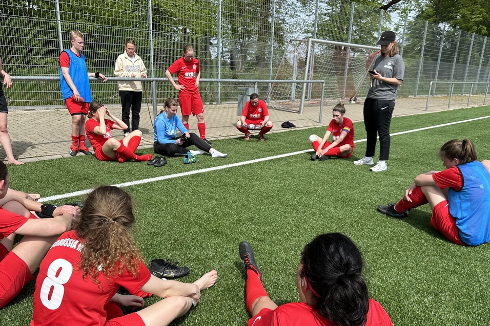 Gruppensitzung mit Fu&szlig;ballspielerinnen in roten Trikots auf einem Kunstrasenplatz, Trainer spricht zu den sitzenden Spielerinnen.