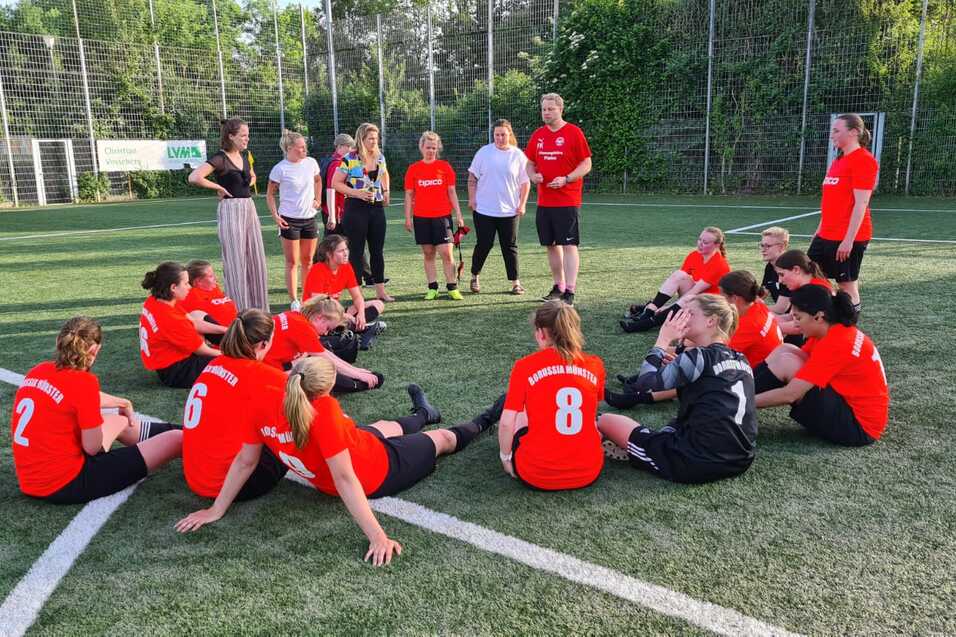 Gruppe von Fu&szlig;ballspielerinnen in roten Trikots sitzt auf dem Rasenfeld, w&auml;hrend Trainer und Betreuerin Anweisungen geben.