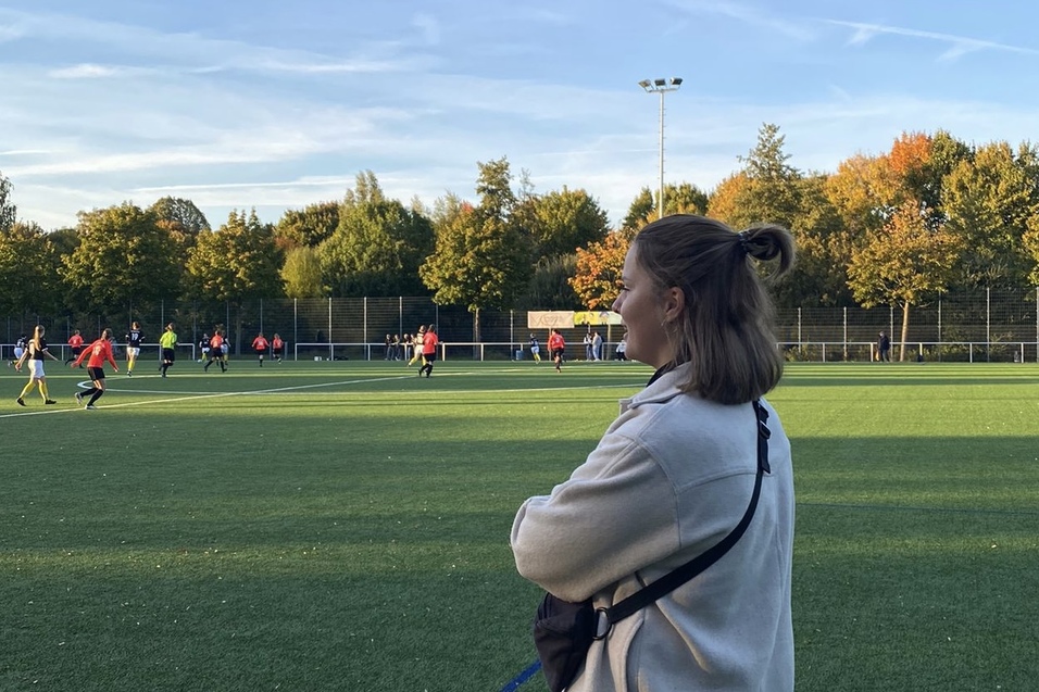 Frau mit kurzem Haar steht auf einem Sportplatz, w&auml;hrend im Hintergrund eine Fu&szlig;ballmannschaft spielt.