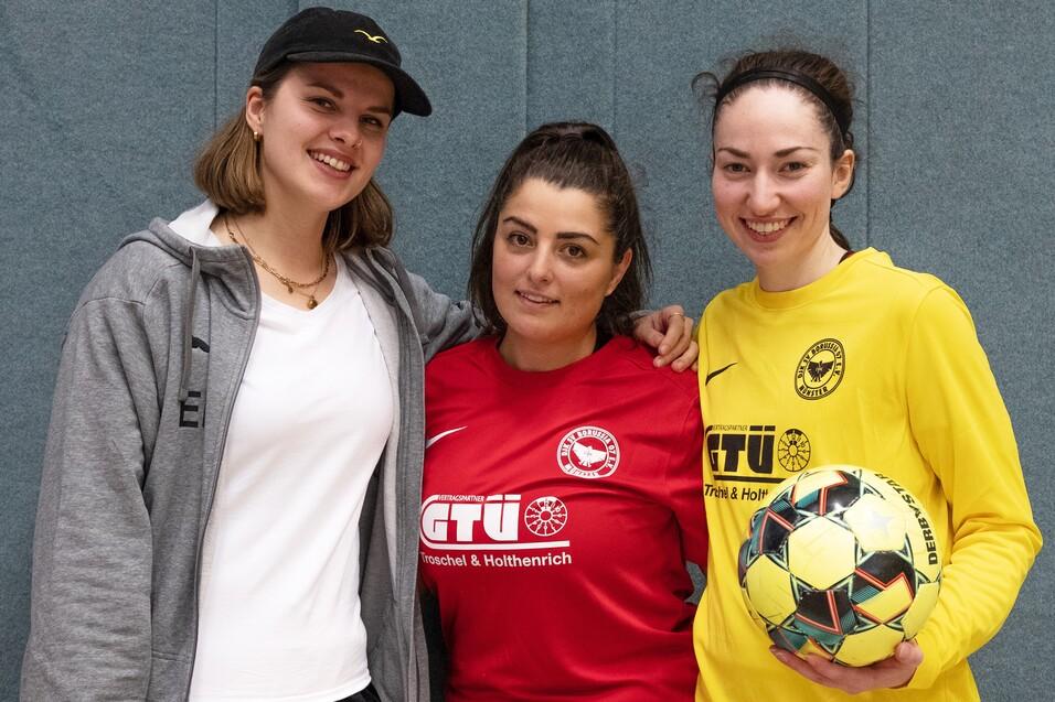 Drei Frauen in Sportkleidung stehen vor einer grauen Wand; eine h&auml;lt einen Fu&szlig;ball in der Hand.