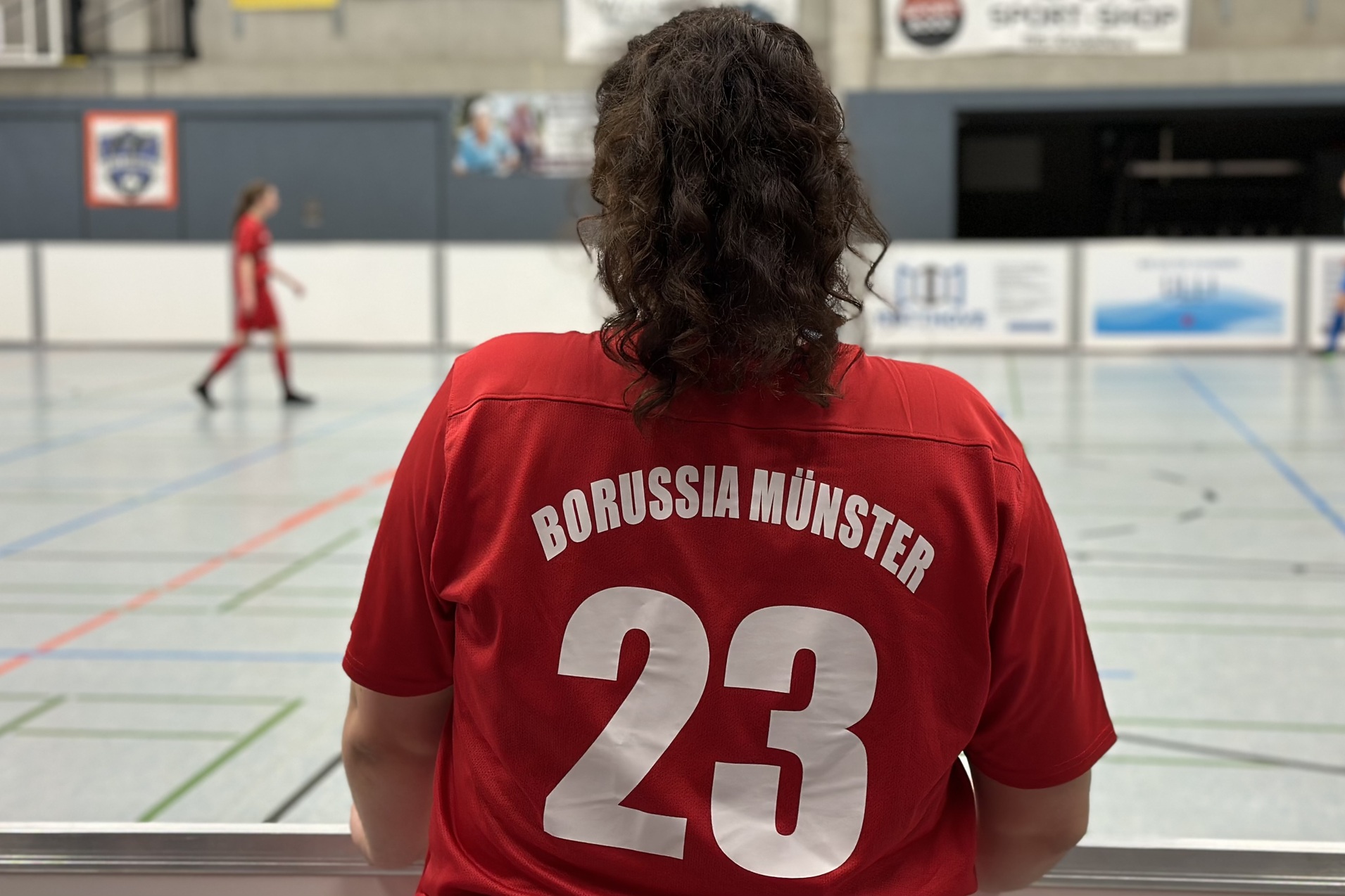 R&uuml;ckansicht einer Person in rotem Trikot mit &bdquo;Borussia M&uuml;nster&ldquo; und der Nummer 23, w&auml;hrend ein Hallenspiel stattfindet.