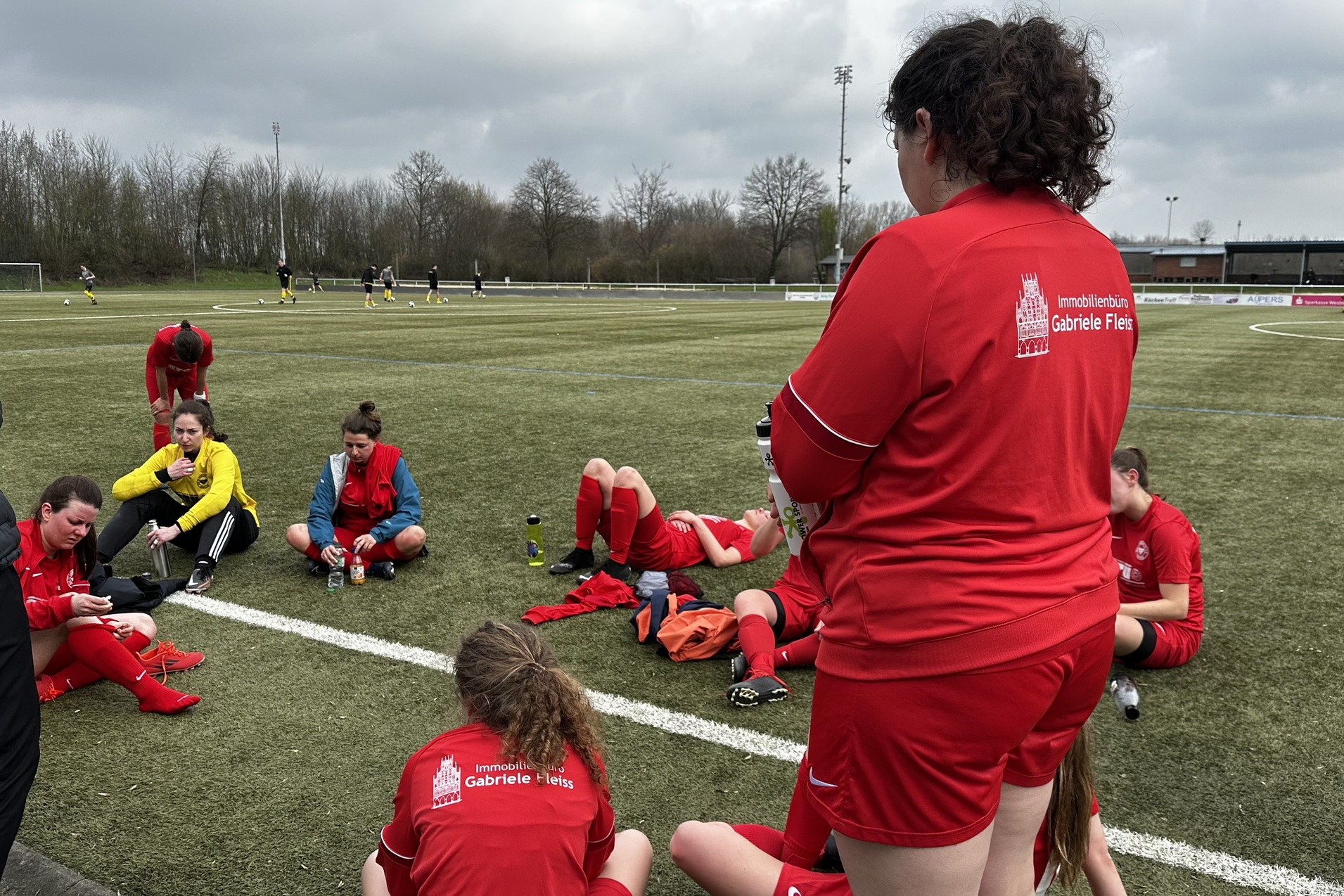 Eine Gruppe von Fu&szlig;ballspielerinnen in roten Trikots sitzt und entspannt sich auf einem Sportplatz mit Kunstrasen.