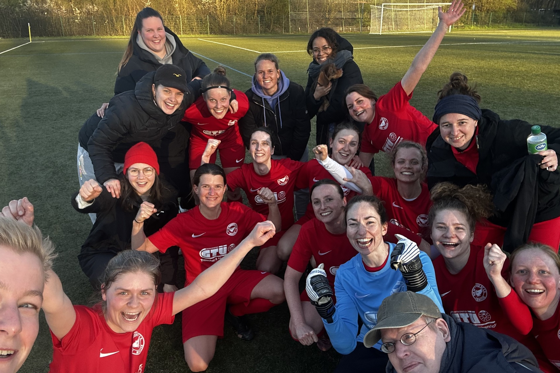 Gruppenselfie von Frauen in roten Fu&szlig;balltrikots auf einem Feld, alle l&auml;cheln und feiern gemeinsam.