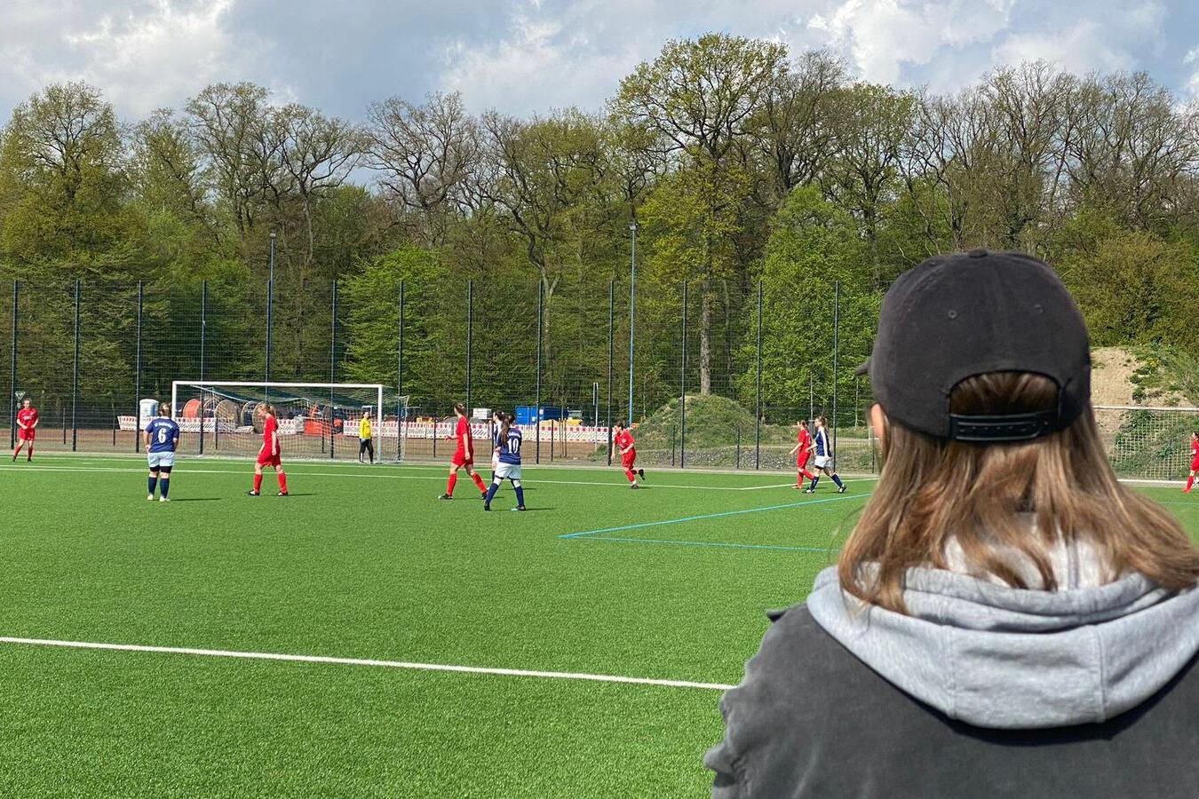 Zuschauer blickt auf ein Fu&szlig;ballspiel zwischen zwei Mannschaften in roten und blauen Trikots auf einem gr&uuml;nen Rasenplatz.