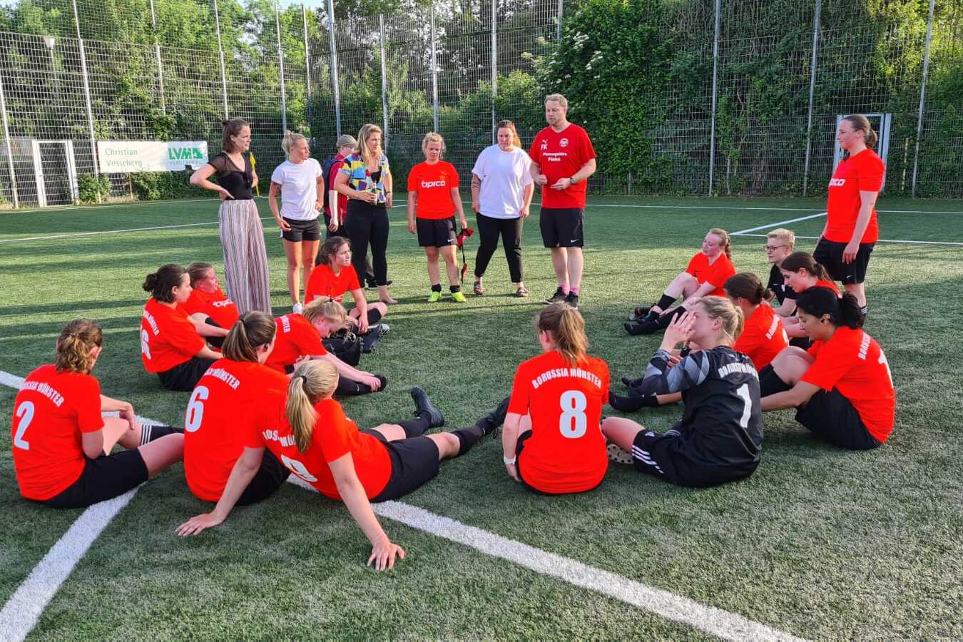 Gruppe von Fu&szlig;ballspielerinnen in roten Trikots sitzt auf dem Rasenfeld, w&auml;hrend Trainer und Betreuerin Anweisungen geben.