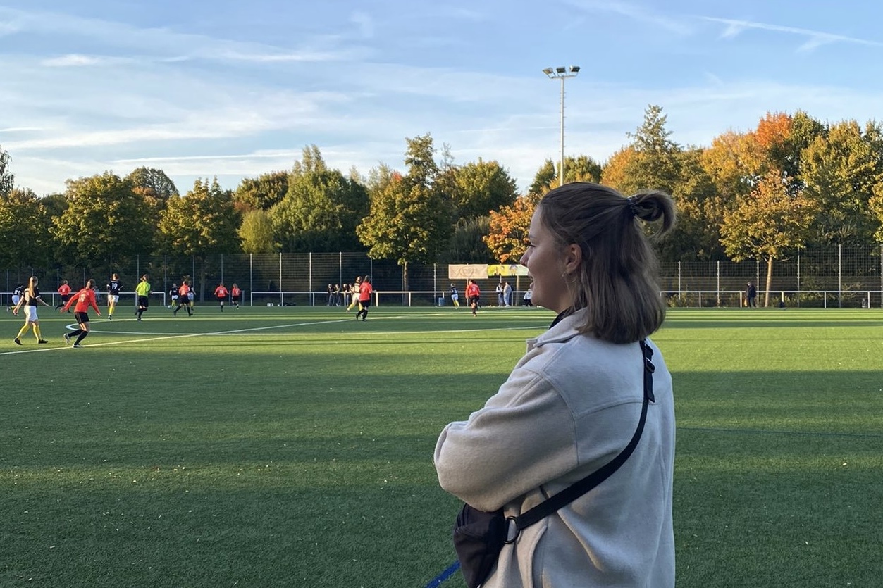 Frau mit kurzem Haar steht auf einem Sportplatz, w&auml;hrend im Hintergrund eine Fu&szlig;ballmannschaft spielt.