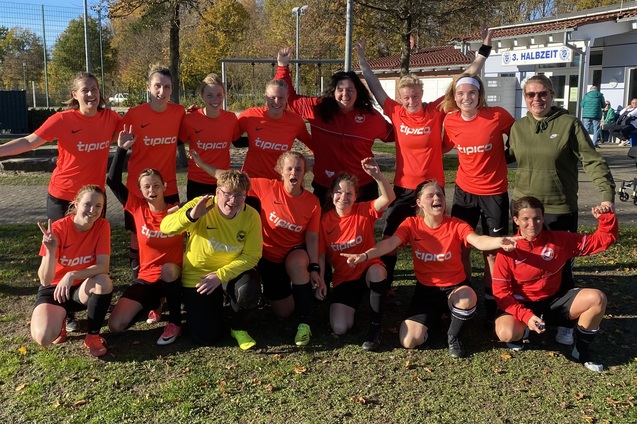 Gruppe von Fu&szlig;ballspielerinnen in roten Trikots posiert fr&ouml;hlich auf einem Sportplatz. Im Hintergrund ein Geb&auml;ude.