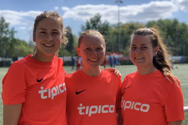 Drei Spielerinnen in orangefarbener Sportkleidung stehen l&auml;chelnd auf einem Fu&szlig;ballplatz mit blauer Himmel im Hintergrund.