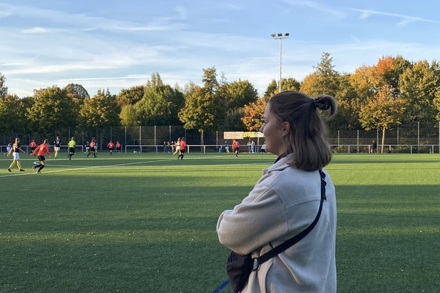 Frau mit kurzem Haar steht auf einem Sportplatz, w&auml;hrend im Hintergrund eine Fu&szlig;ballmannschaft spielt.