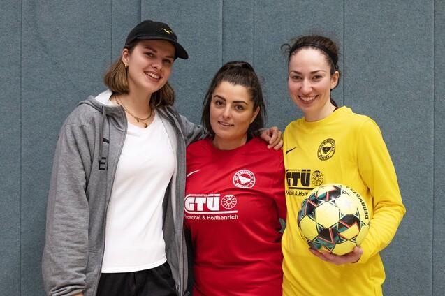 Drei Frauen in Sportkleidung stehen vor einer grauen Wand; eine h&auml;lt einen Fu&szlig;ball in der Hand.