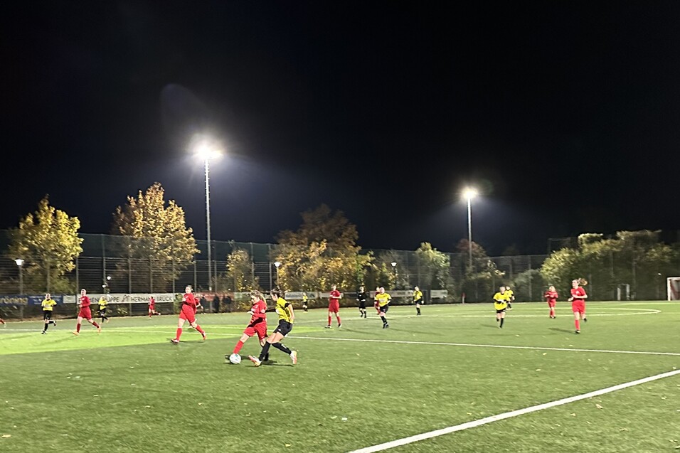 Fu&szlig;ballspiel bei Nacht mit Spielern in roten und gelben Trikots auf einem Kunstrasenplatz unter Flutlicht.