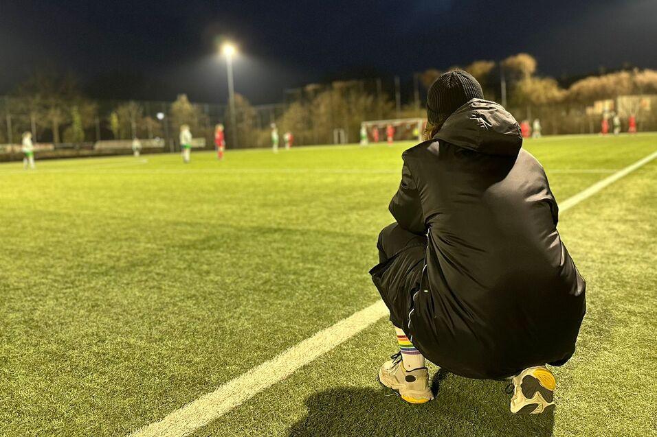 Person im schwarzen Mantel hockt am Rand eines Fu&szlig;ballfeldes, w&auml;hrend im Hintergrund ein Spiel stattfindet.