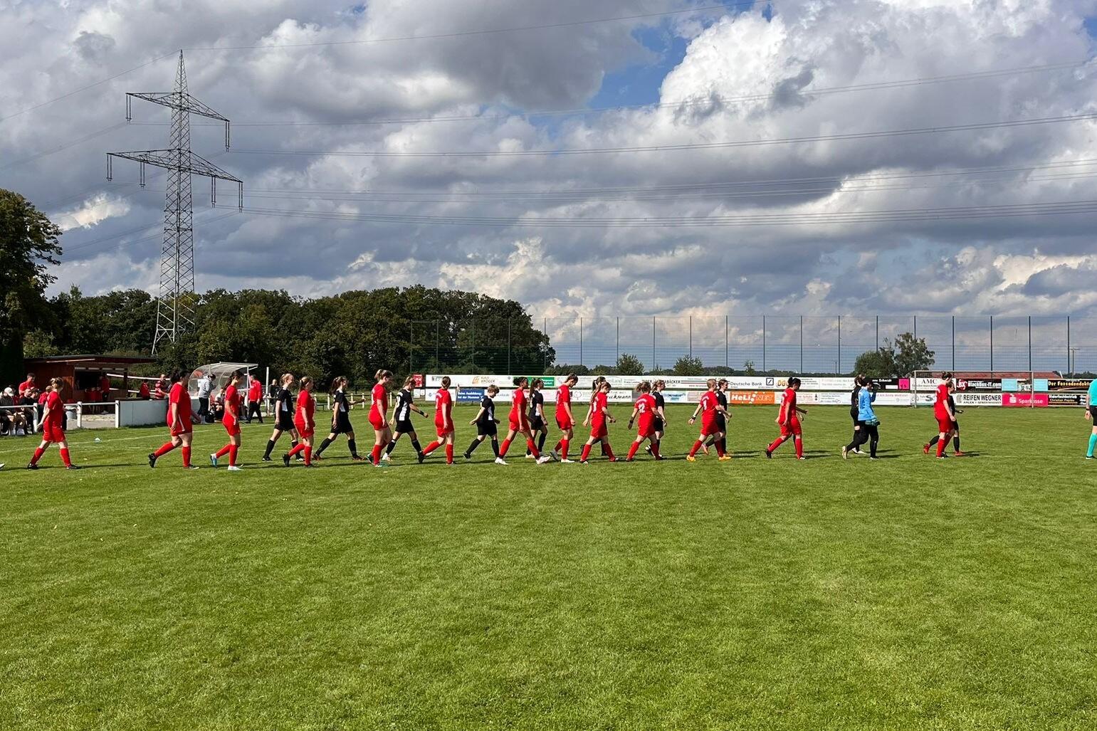 Fu&szlig;ballmannschaften in roten Trikots marschieren auf einem gr&uuml;nen Platz unter einem bew&ouml;lkten Himmel zum Spielbeginn.