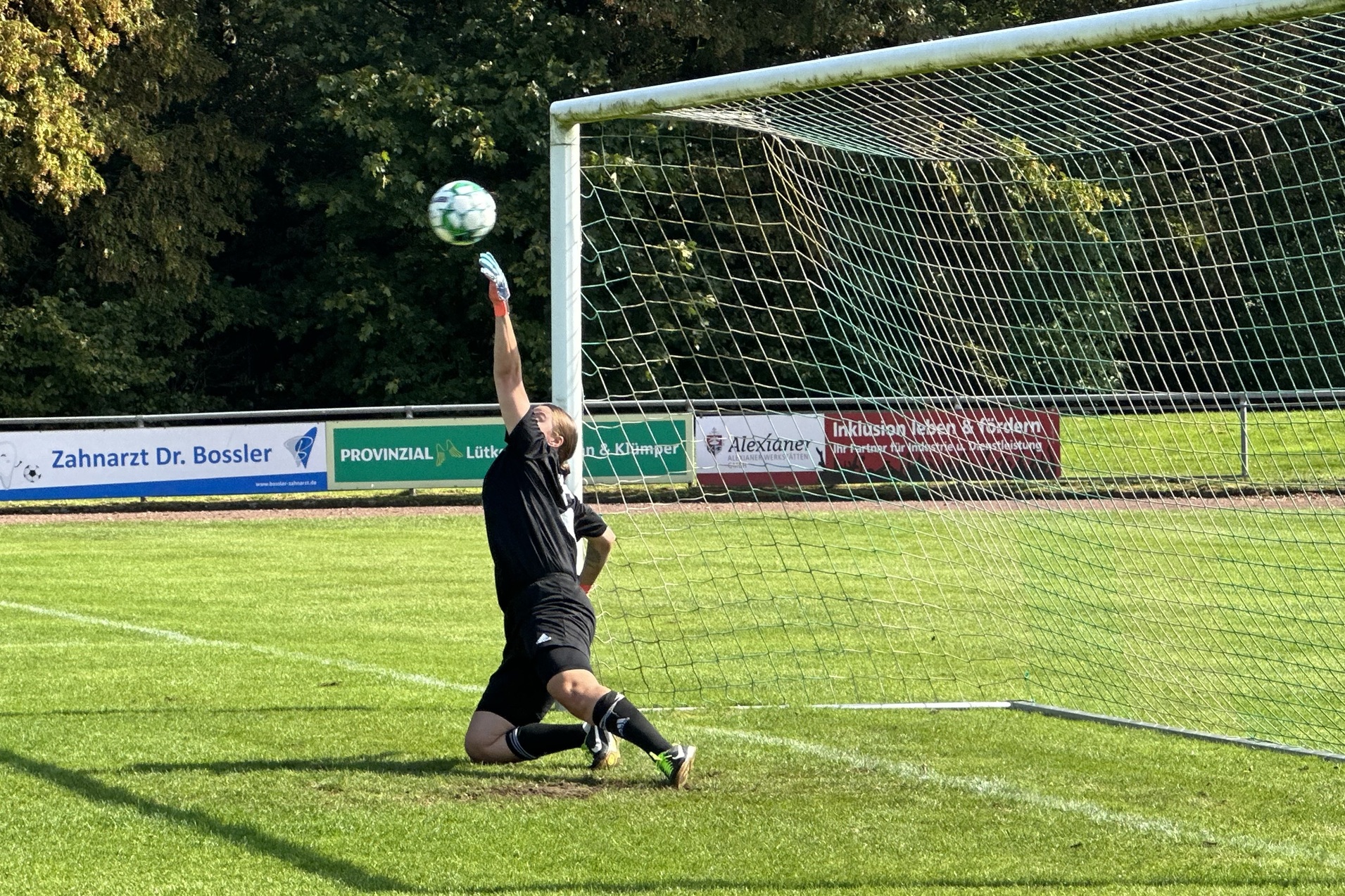 Torwart springt auf dem Fu&szlig;ballfeld, um einen Ball im h&ouml;heren Bogen zu halten, w&auml;hrend er vor dem Tor kniet.