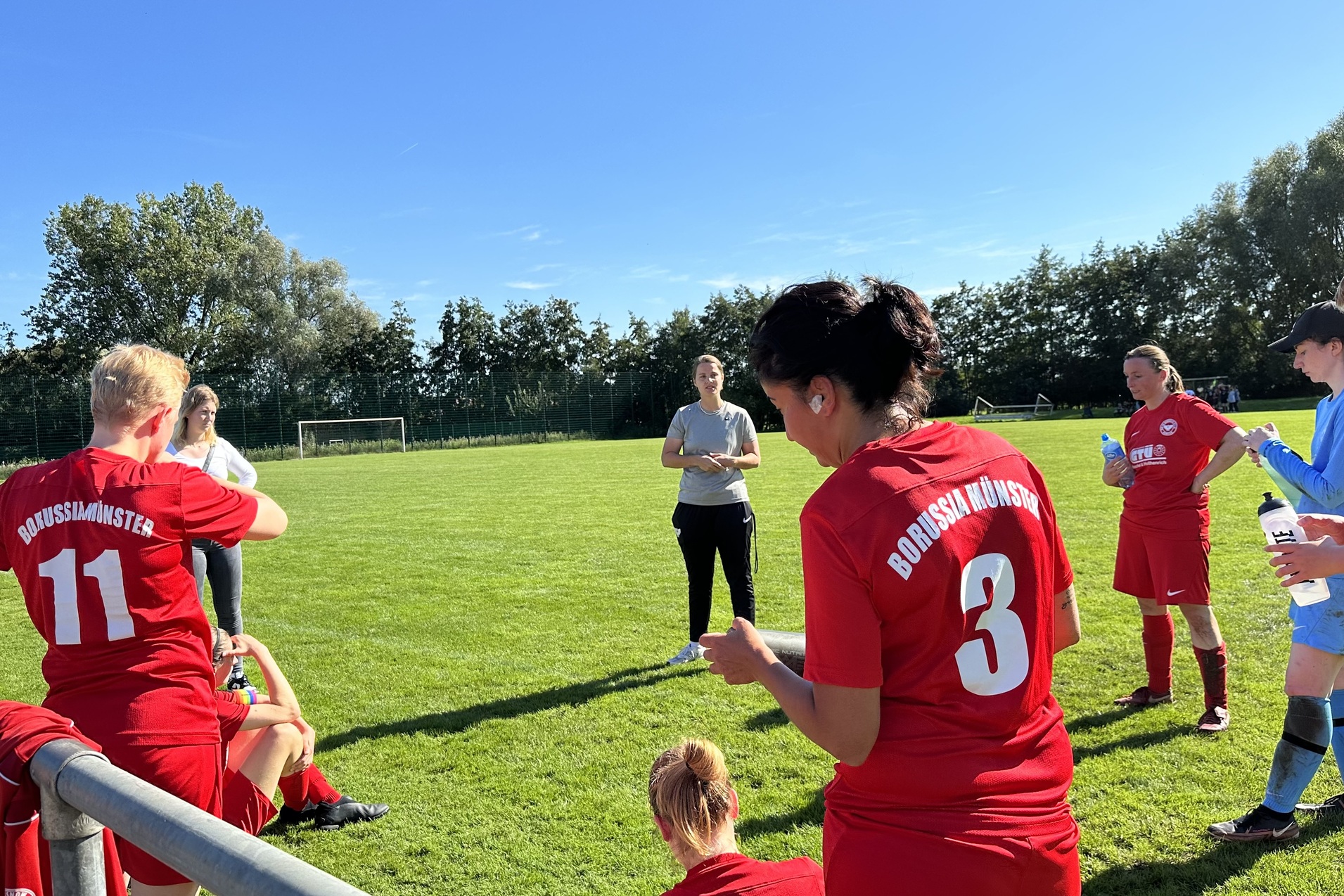 Fu&szlig;ballmannschaft in roten Trikots versammelt sich um eine Trainerin auf dem gr&uuml;nen Platz bei klarem Himmel.