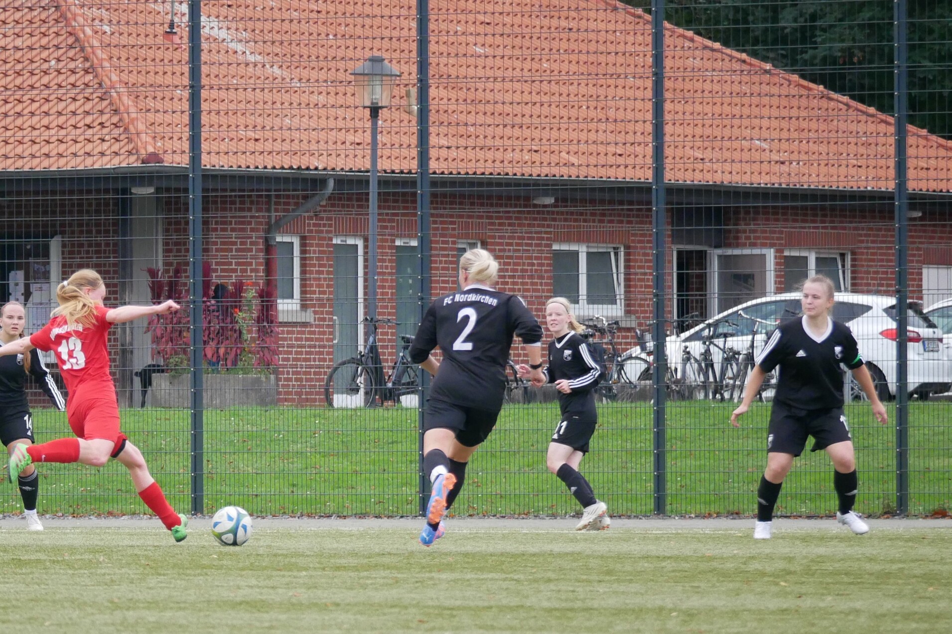 Eine Spielerin in rotem Trikot schie&szlig;t einen Ball auf einem Fu&szlig;ballplatz, umgeben von Gegnerinnen in Schwarz.