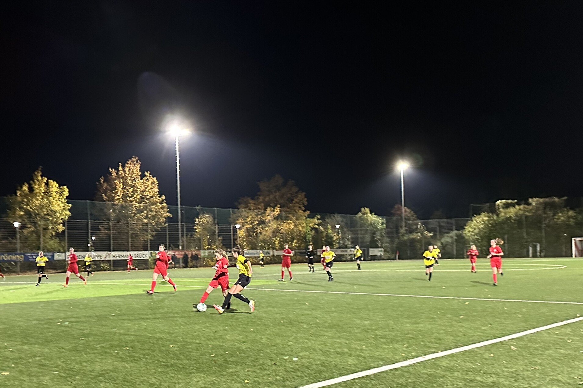 Fu&szlig;ballspiel bei Nacht mit Spielern in roten und gelben Trikots auf einem Kunstrasenplatz unter Flutlicht.