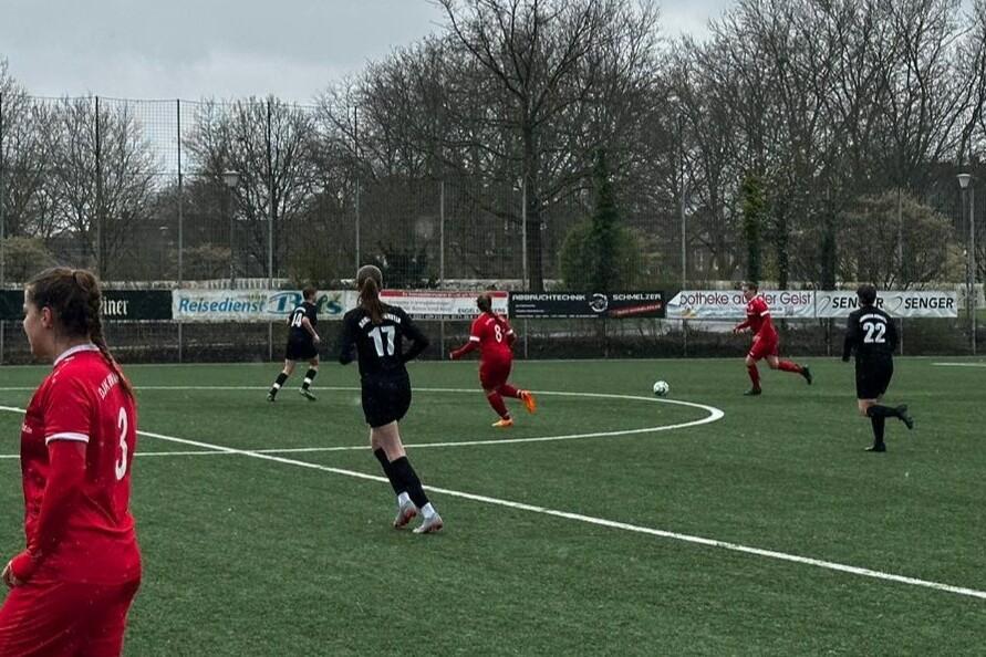 Spielszene auf einem Fu&szlig;ballplatz mit Spielerinnen in roten und schwarzen Trikots, die einem Ball nachlaufen.
